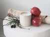 Makana concrete candle on rustic white stool with two fresh red apples and white birch branch in background