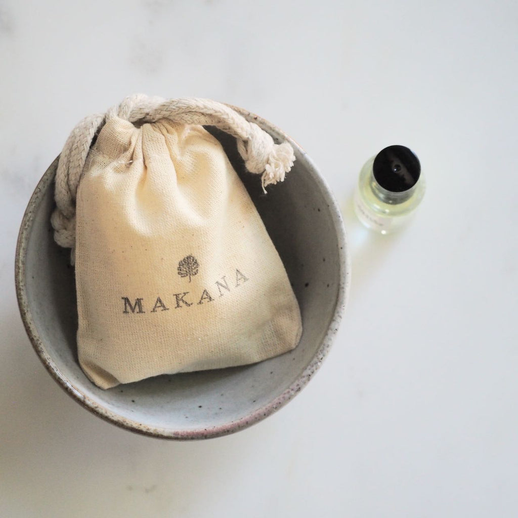 Makaka Perfume on white marble next to artisan bowl that is holding muslin pouch with the name Makana stamped on it