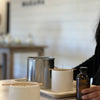 Concrete candle in foreground sitting in front of silver pour pot and another white concrete vessel. Woman's hand holding amber fragrance bottle. Makana logo blurry in background on white shiplap wall.
