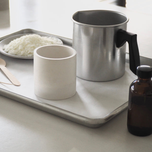 White concrete vessel with silver pour pot sitting on a silver metal tray. Amber fragrance bottle in the foreground and coconut  soy wax flakes in the background.